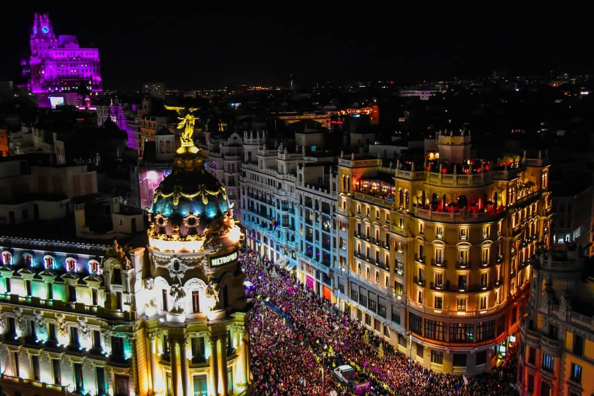Nächtliche Luftaufnahme einer belebten, mit Lichtern geschmückten Straße im Zentrum von Madrid, gefüllt mit Menschen und festlicher Atmosphäre, mit den ikonischen Gebäuden der Stadt im Hintergrund.