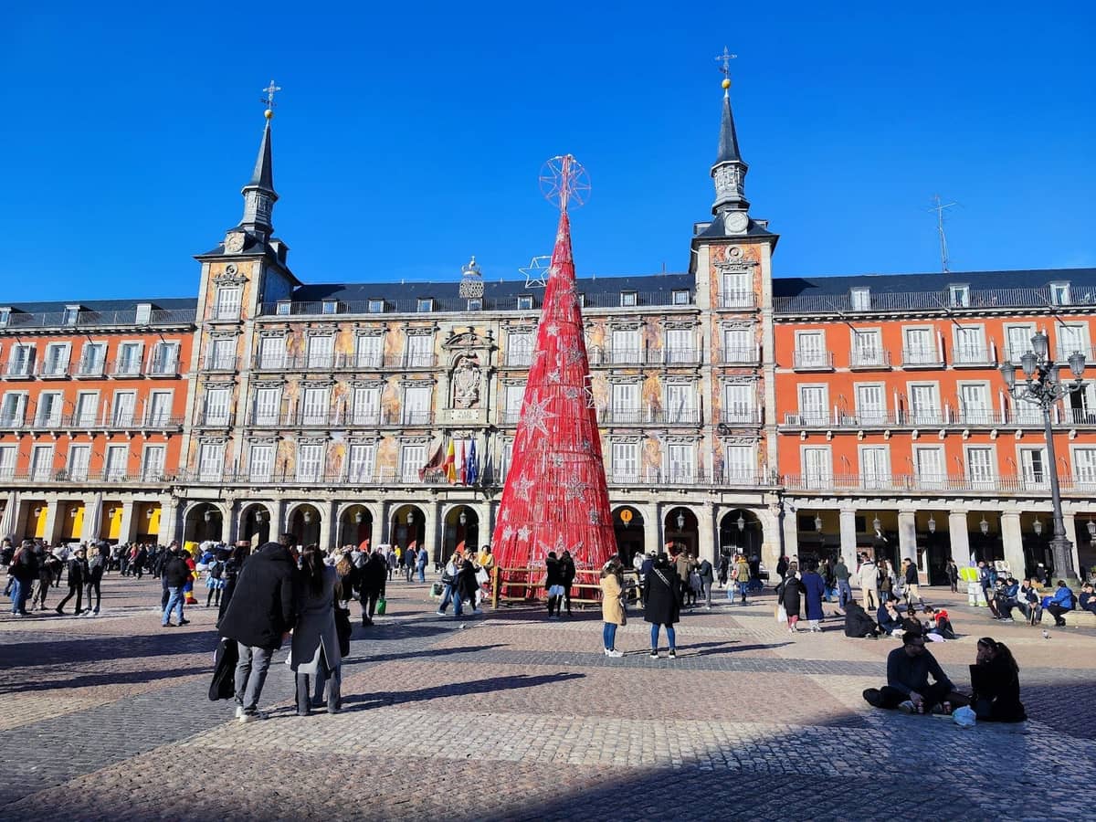 Menschen versammeln sich auf der Plaza Mayor in Madrid, die an einem sonnigen Wintertag mit einem großen roten Weihnachtsbaum in der Mitte geschmückt ist.