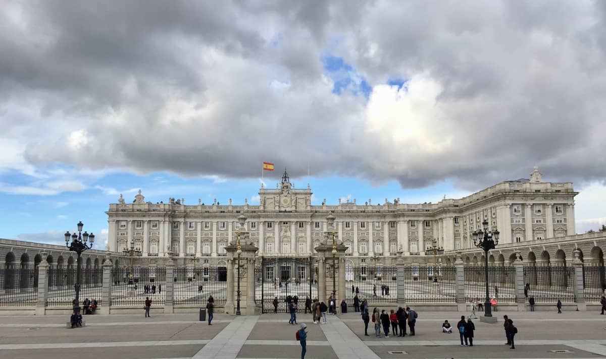 Weitwinkelansicht des Königspalastes von Madrid mit Menschen auf dem Platz, unter dramatischen Winterwolken und einer spanischen Flagge auf dem Gebäude.