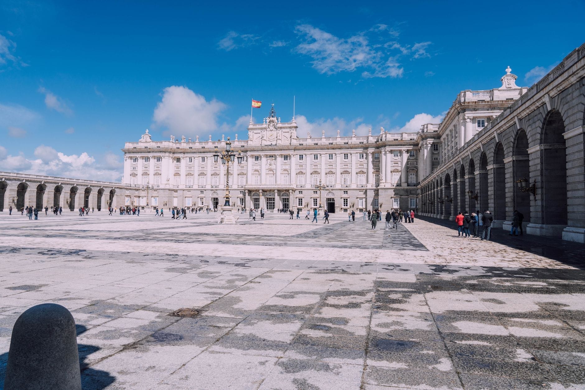 Vista ampla do Palácio Real de Madrid com uma praça espaçosa e ensolarada, alguns visitantes caminhando e um céu azul vibrante ao fundo.