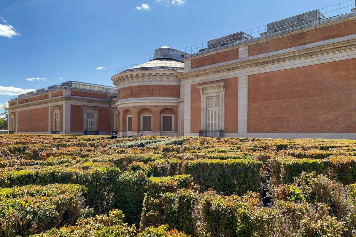 Exterior view of the Prado Museum with its red brick facade and neatly trimmed hedges in the foreground, under a blue sky.