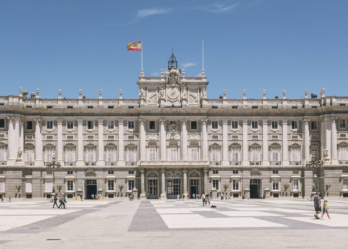 Wide view of the Royal Palace of Madrid with the Spanish flag flying and people walking in the courtyard.