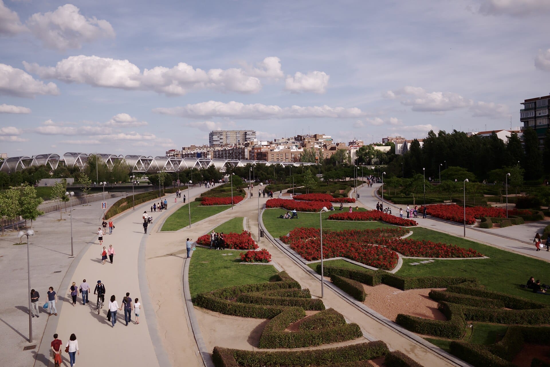 Ampia veduta del parco Madrid Río con giardini paesaggistici, passerella moderna, percorsi pedonali e persone che godono di un pomeriggio di sole all'aperto.