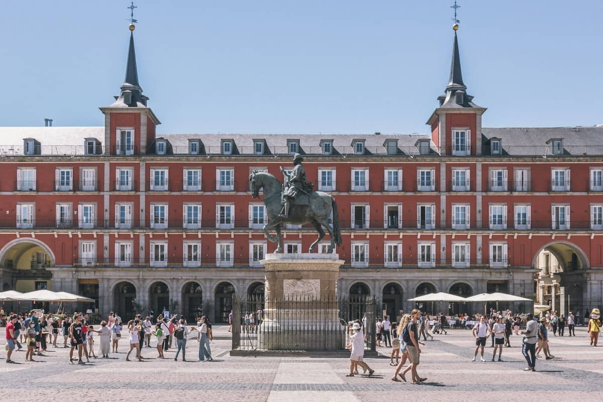 Scène animée sur la Plaza Mayor à Madrid avec des touristes se promenant et la statue équestre au centre, bâtiments historiques en arrière-plan.
