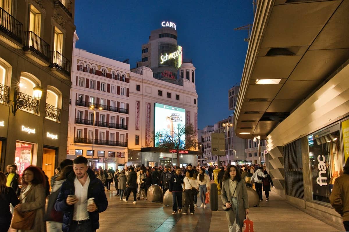 Rue animée du centre de Madrid au crépuscule, avec des passants, des boutiques et des devantures illuminées, évoquant l'atmosphère d'un quartier urbain animé.