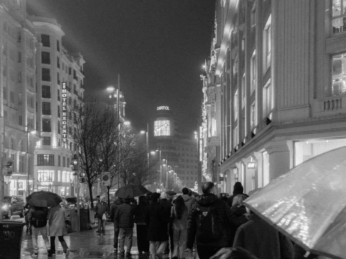 Rue animée de Madrid la nuit avec des personnes tenant des parapluies, un pavé brillant mouillé et des bâtiments emblématiques visibles en arrière-plan.