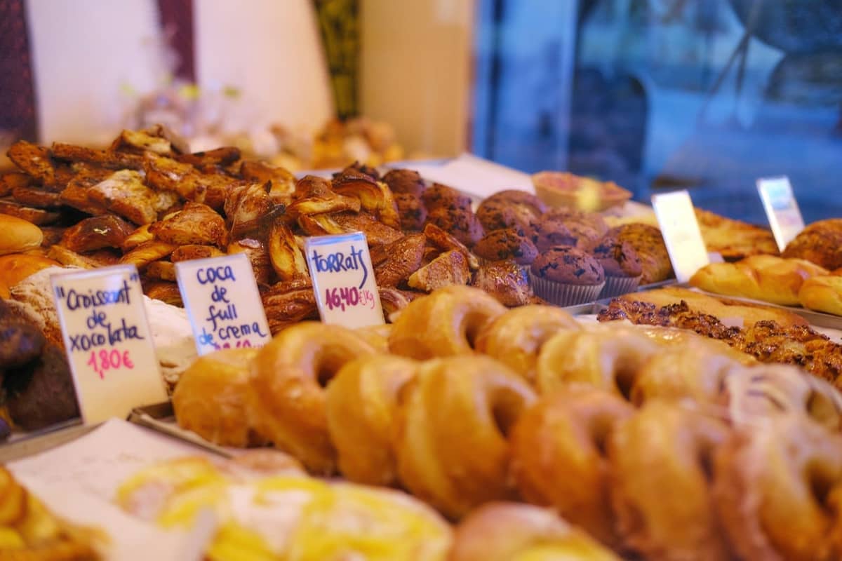 Nahaufnahme einer Bäckerei-Auslage mit verschiedenen traditionellen Gebäckstücken, die mit spanischen Namen und Preisen beschriftet sind, durch ein Schaufenster betrachtet.