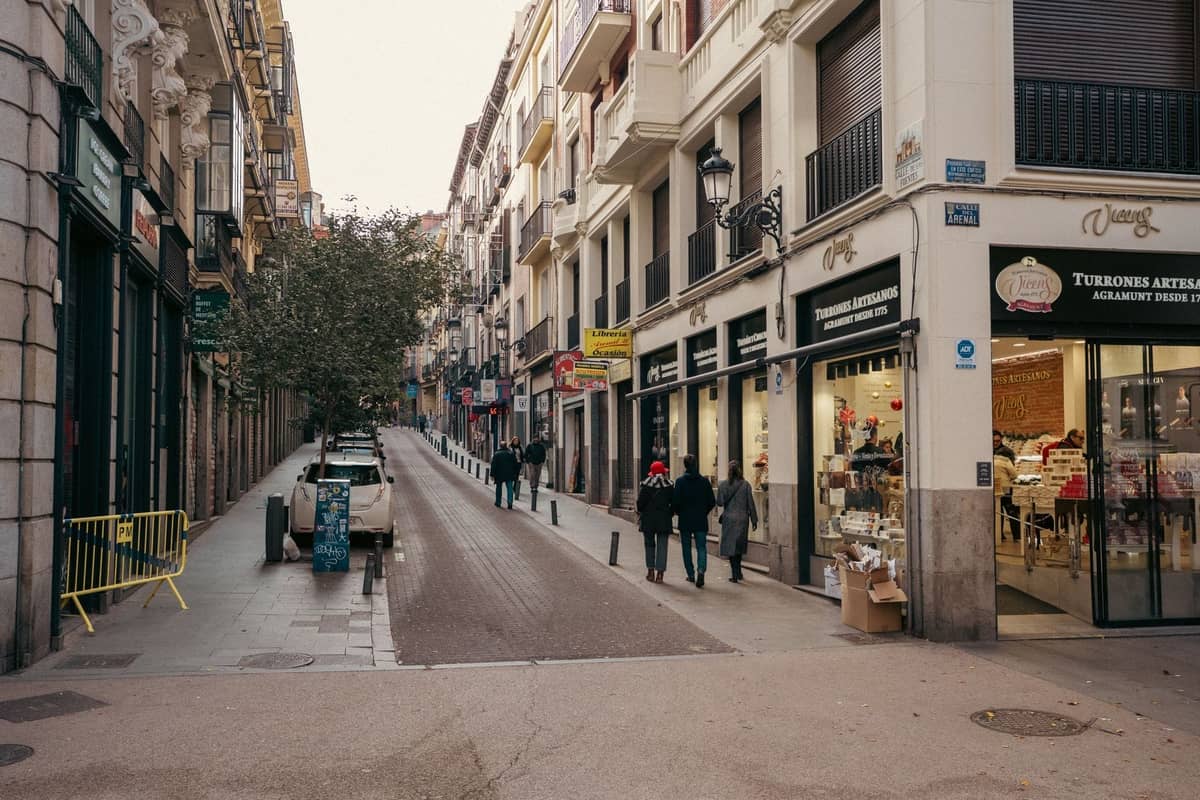 Calle comercial estrecha en Madrid con personas caminando, flanqueada por edificios modernos y antiguos con diversas tiendas y cafés.
