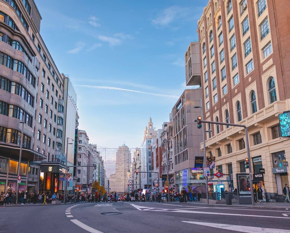 Vista amplia de la Gran Vía en Madrid, con compradores, tráfico y edificios clásicos a ambos lados de esta concurrida calle comercial bajo un cielo azul despejado.