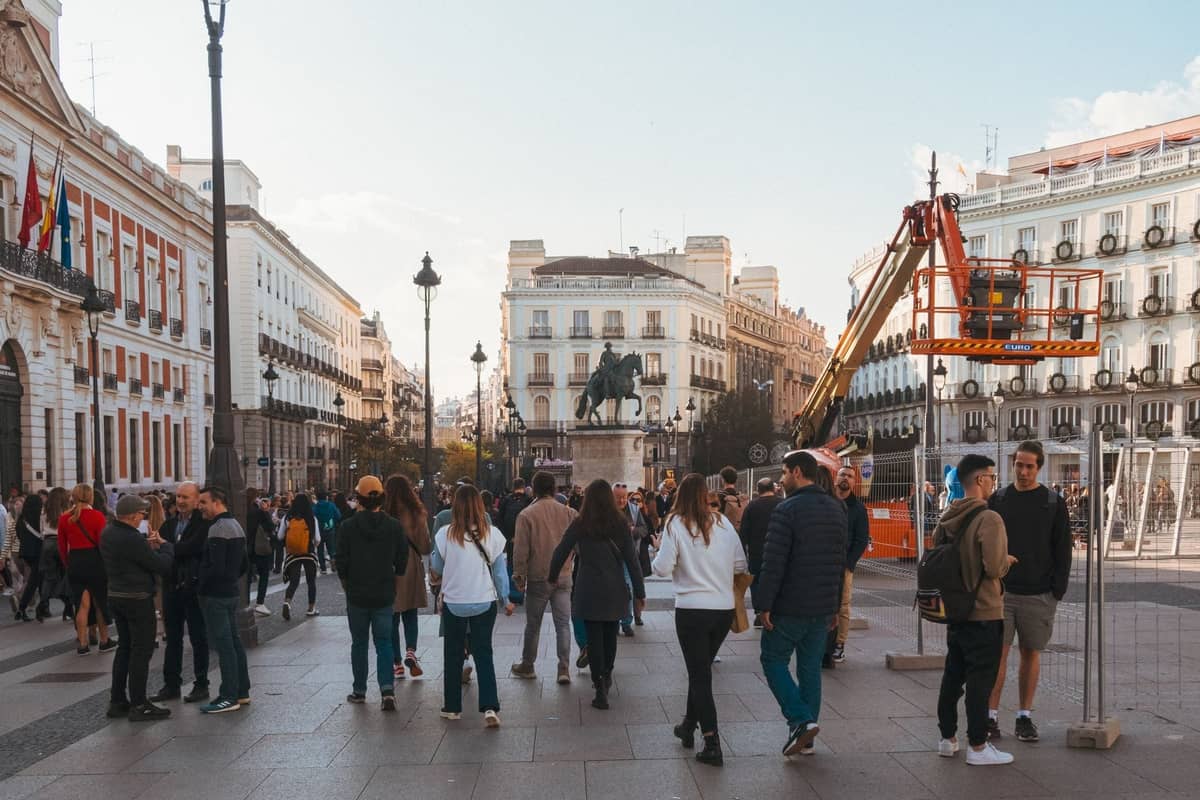 Madrid'in merkezinde yürüyen insan grupları, tarihi binalar ve arka planda bir heykel ile kalabalık bir meydan, gün ışığında.