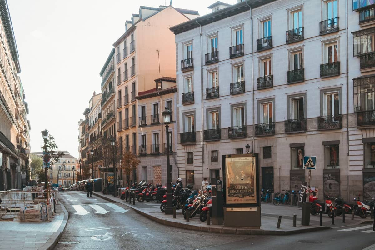 Madrid'in klasik konut binalarıyla çevrili, kaldırımda park etmiş scooter'ların ve birkaç yayaın bulunduğu sakin bir sokağının geniş görünümü.