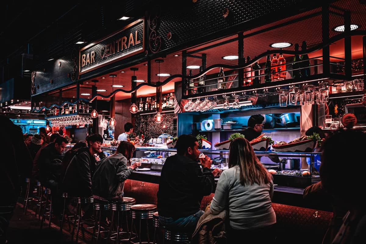 Belebte Tapas-Bar in Madrid mit Gästen, die an einer Theke sitzen und Getränke und Tapas genießen, Bar Central-Schild darüber, lebhafte Abendatmosphäre.