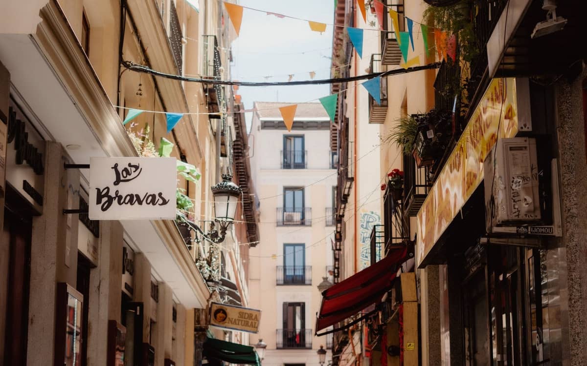 Eine enge Straße in Madrid, gesäumt von bunten Barschildern, darunter 'Las Bravas', Balkonen und festlichen hängenden Fahnen, die ein lebhaftes Tapas-Viertel heraufbeschwören.