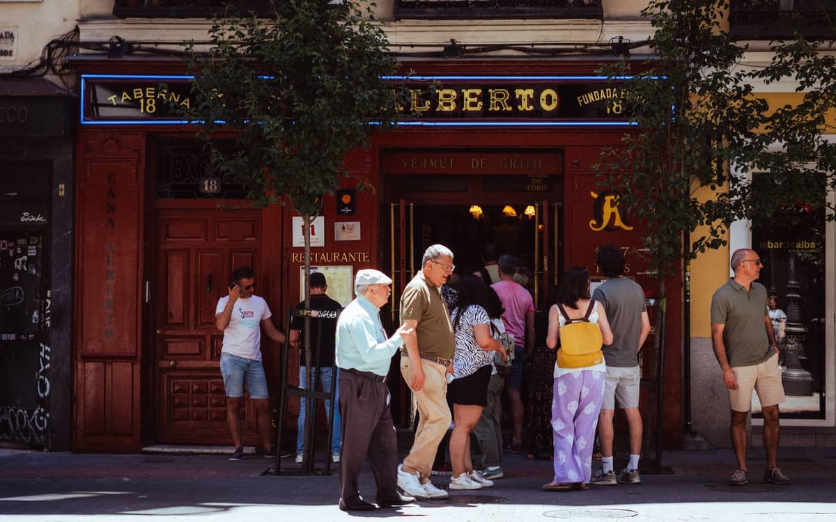 Menschen vor dem Eingang einer klassischen Bar in Madrid an einem sonnigen Tag, mit Tavernenschild und Straßenleben im Hintergrund.