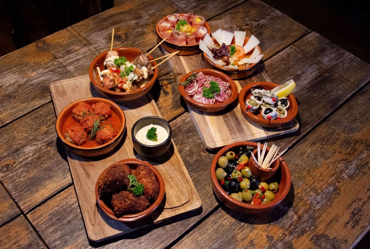 Ein Holztisch mit einer Auswahl klassischer spanischer Tapas in Tonschalen, darunter Oliven, Kroketten, Käse, Aufschnitt und Meeresfrüchte.