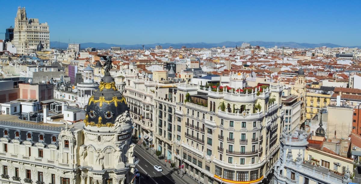 Vista aérea luminosa de la Gran Vía de Madrid y el edificio Metrópolis en un día soleado con cielos azules despejados y montañas al fondo.