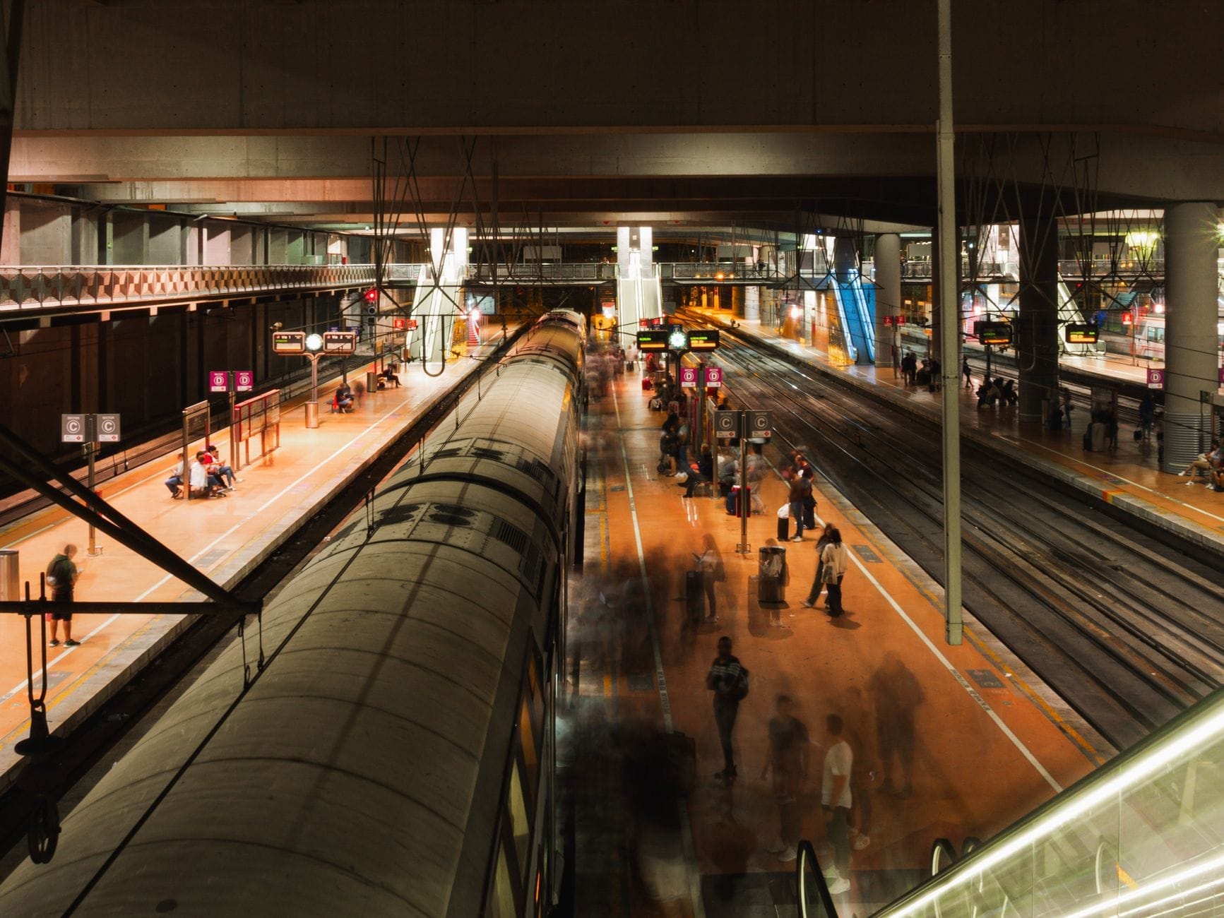 Uma movimentada estação de trem com viajantes caminhando na plataforma e trens em vários trilhos sob uma iluminação aconchegante.