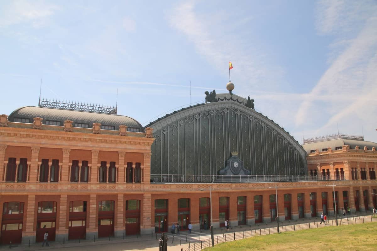 La fachada de ladrillo rojo y cristal de la estación de tren Atocha de Madrid bajo un cielo azul, con una bandera española ondeando en lo alto.