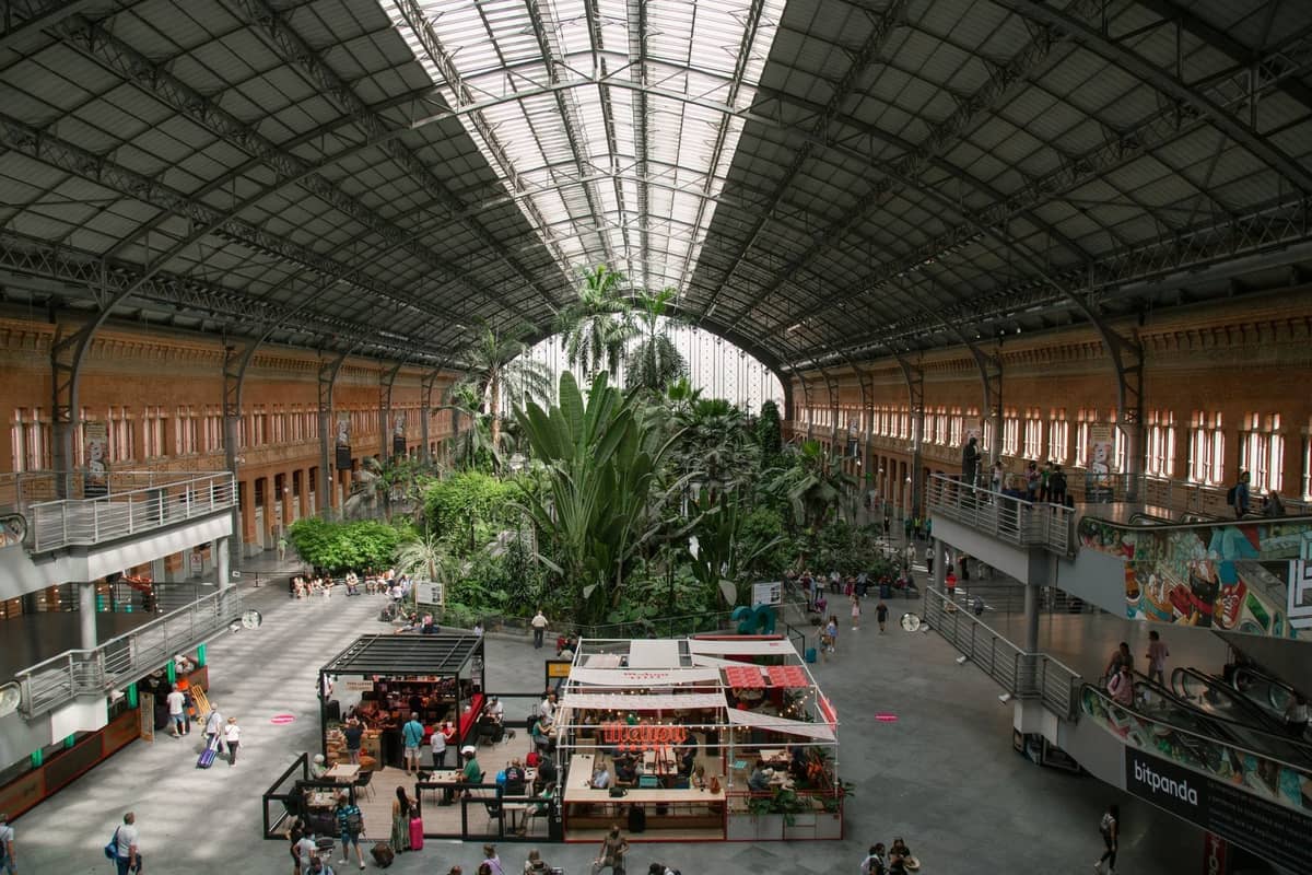Inneres des Bahnhofs Atocha in Madrid mit seinem markanten Glasdach, tropischen Garten und Menschen, die durch die geräumige Halle gehen oder sitzen.