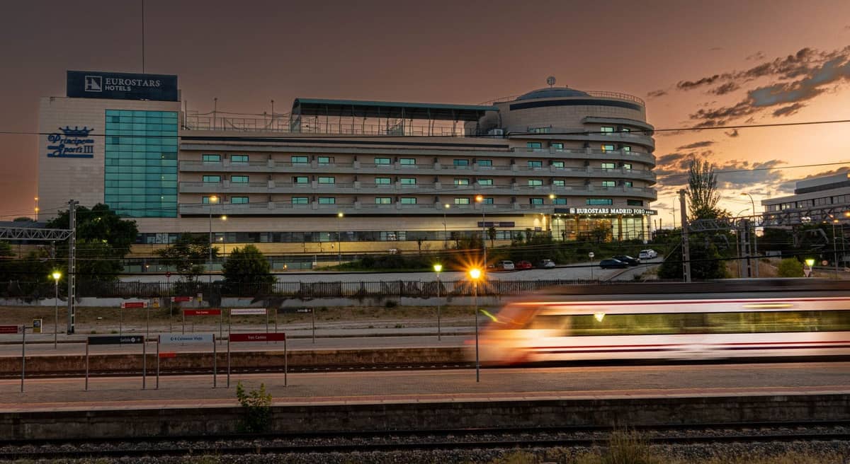 Verschwommener Zug verlässt den Bahnhof Madrid Atocha in der Dämmerung mit Hotel und Parkplatz im Hintergrund