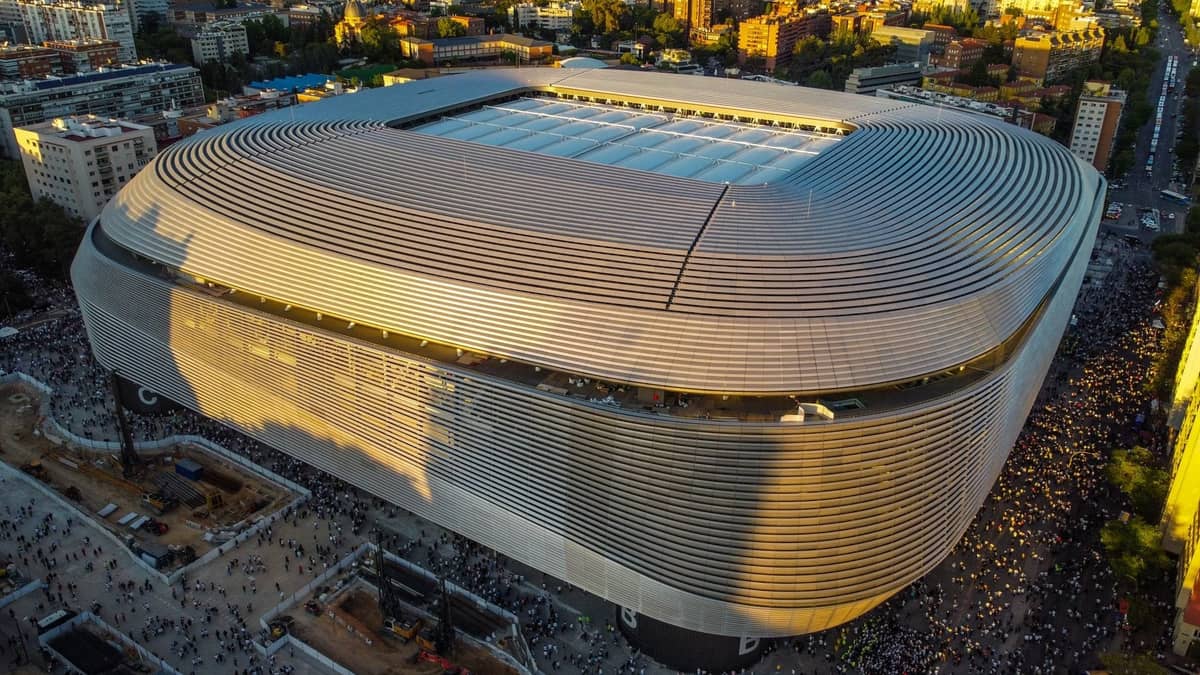 Vista aerea dello Stadio Santiago Bernabéu a Madrid, gremito di tifosi con lo skyline della città sullo sfondo. L'architettura moderna e l'atmosfera calcistica sono chiaramente visibili.