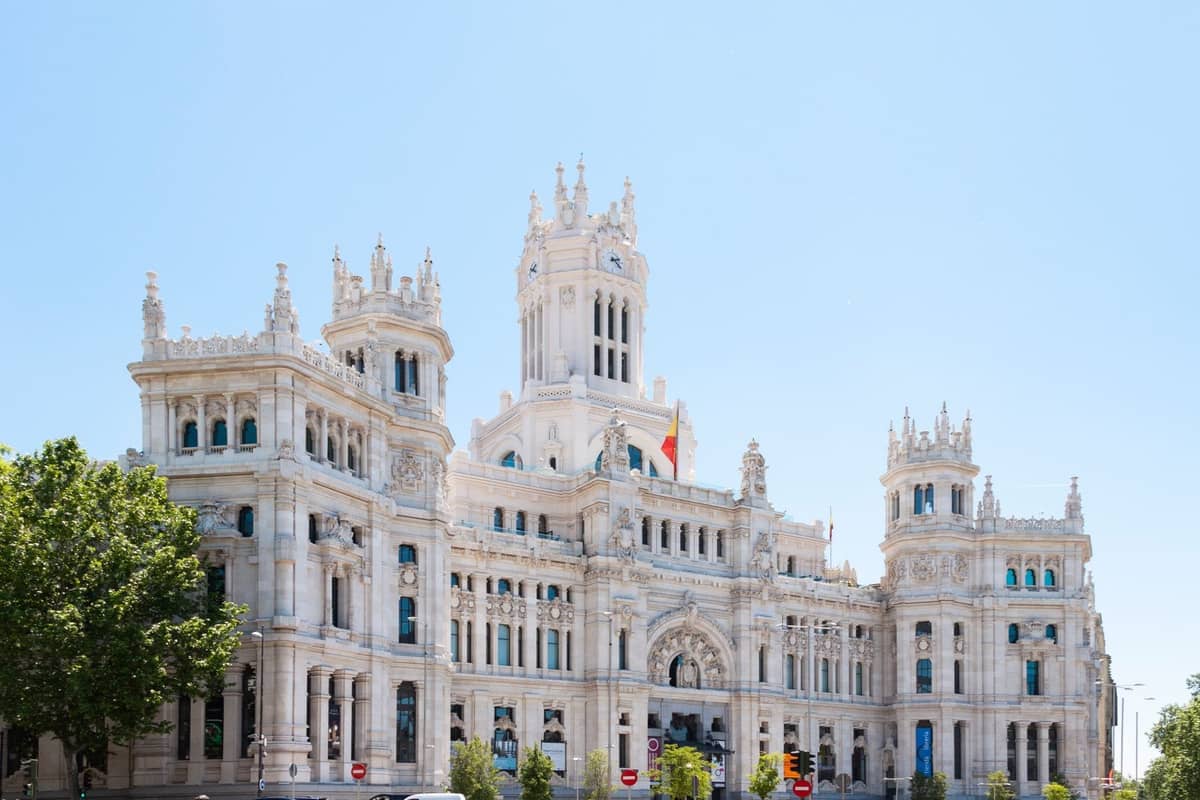 La sontuosa facciata bianca del Palacio de Cibeles di Madrid sotto un cielo azzurro brillante, con bandiere spagnole al vento e alberi verdi di fronte.