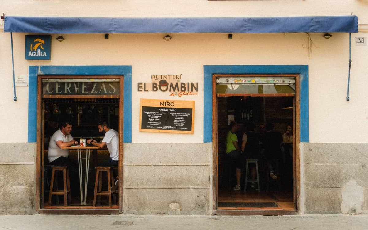 Vista stradale di un bar informale a Madrid con persone sedute a tavoli alti vicino a porte aperte, cornici blu e un'insegna con la scritta El Bombín.