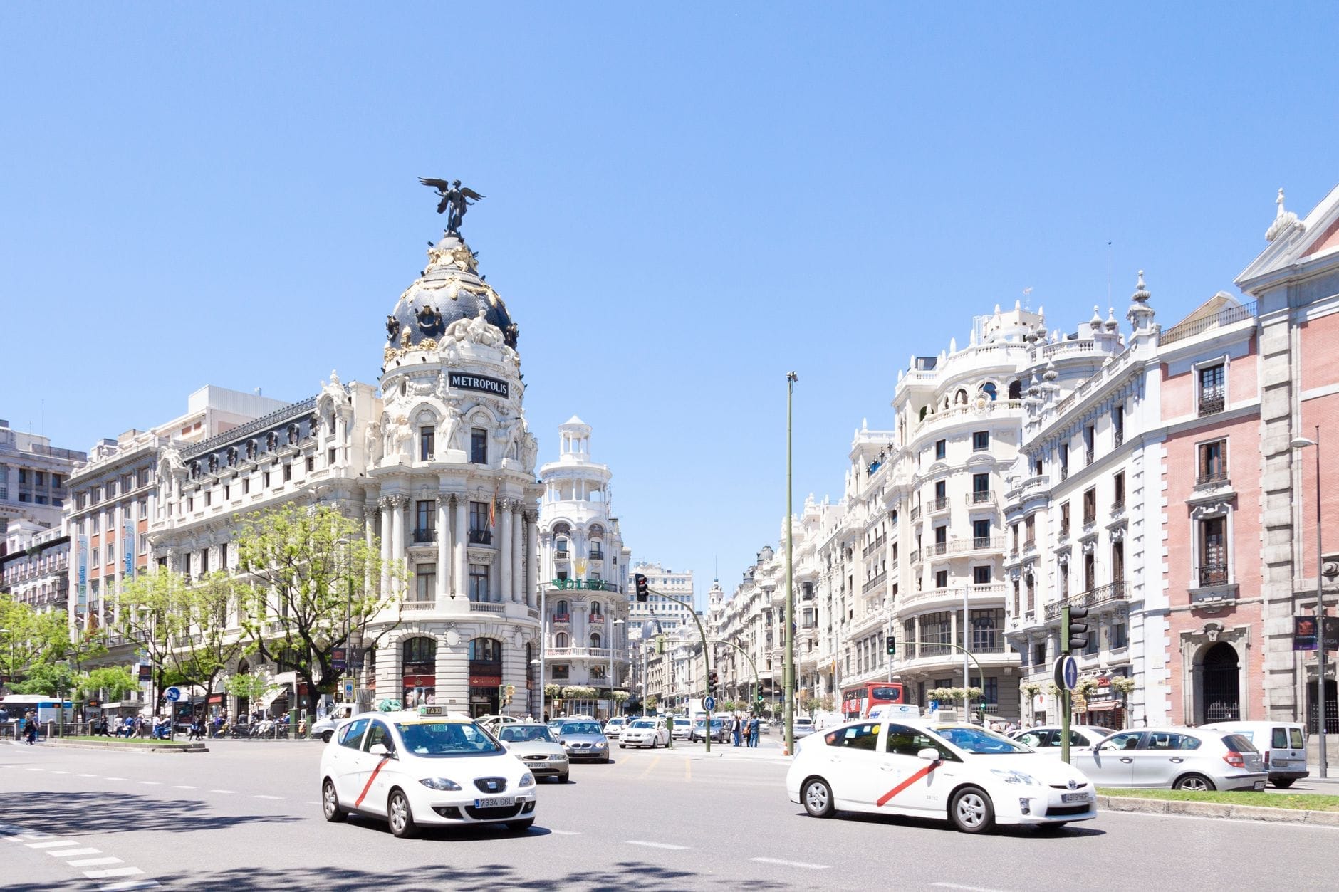 Vista ampla da icônica Gran Via de Madrid com prédios clássicos, trânsito intenso e céu azul, capturando a vida urbana vibrante e a arquitetura histórica da cidade.