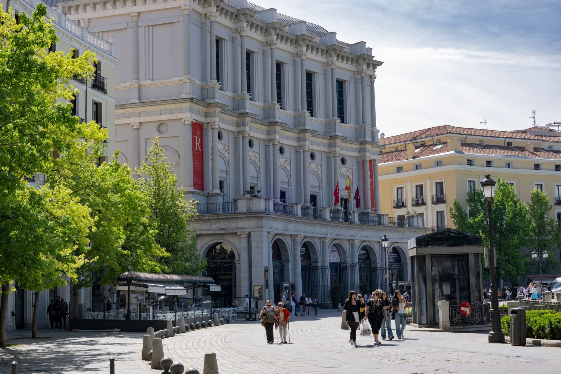 Pessoas passeando perto do elegante Teatro Real de Madri em um dia ensolarado, com árvores verdes e prédios clássicos ao fundo.