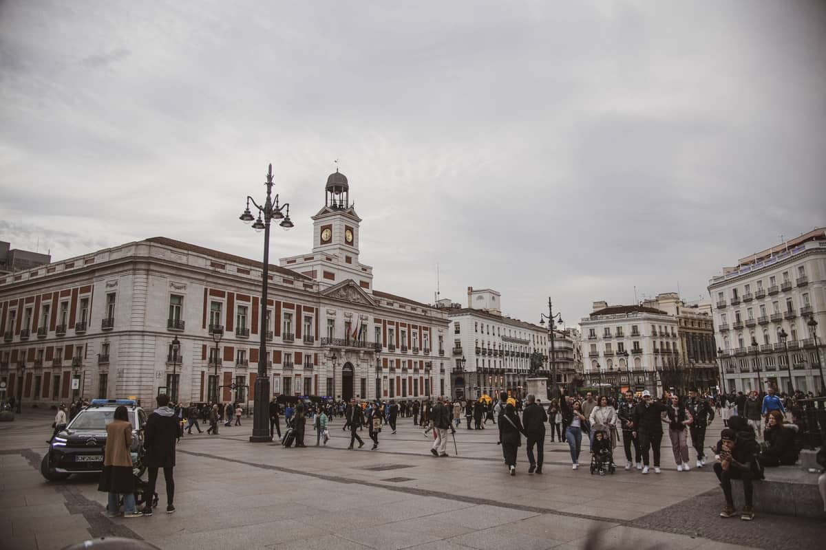 Weitwinkelansicht des Platzes Puerta del Sol in Madrid mit Menschenmassen, historischen Gebäuden und dem berühmten Uhrenturm im Hintergrund.