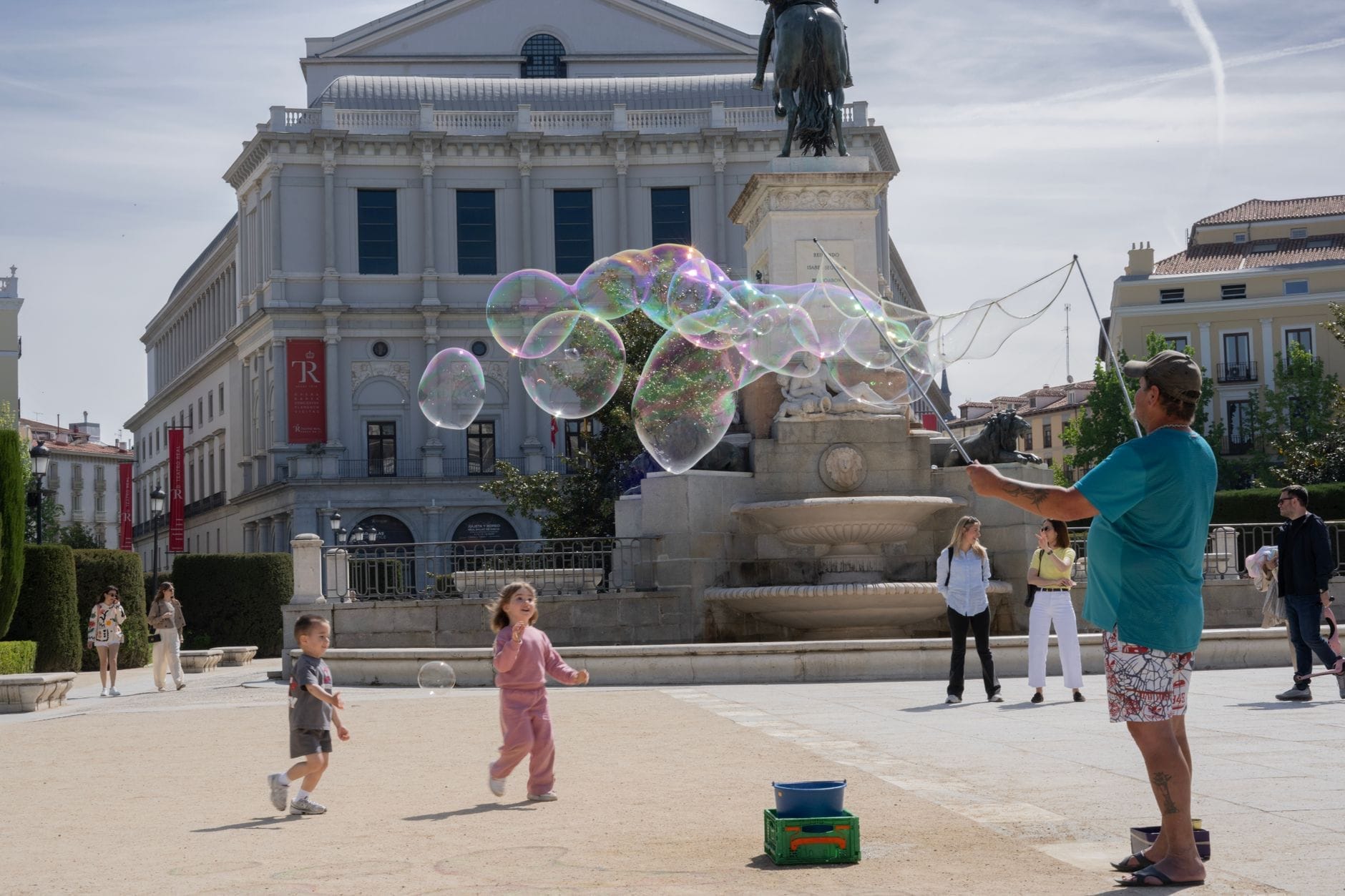 Crianças brincando animadas com bolhas de sabão gigantes em uma praça ensolarada de Madrid, com um prédio imponente e uma fonte ao fundo — diversão em família pura.