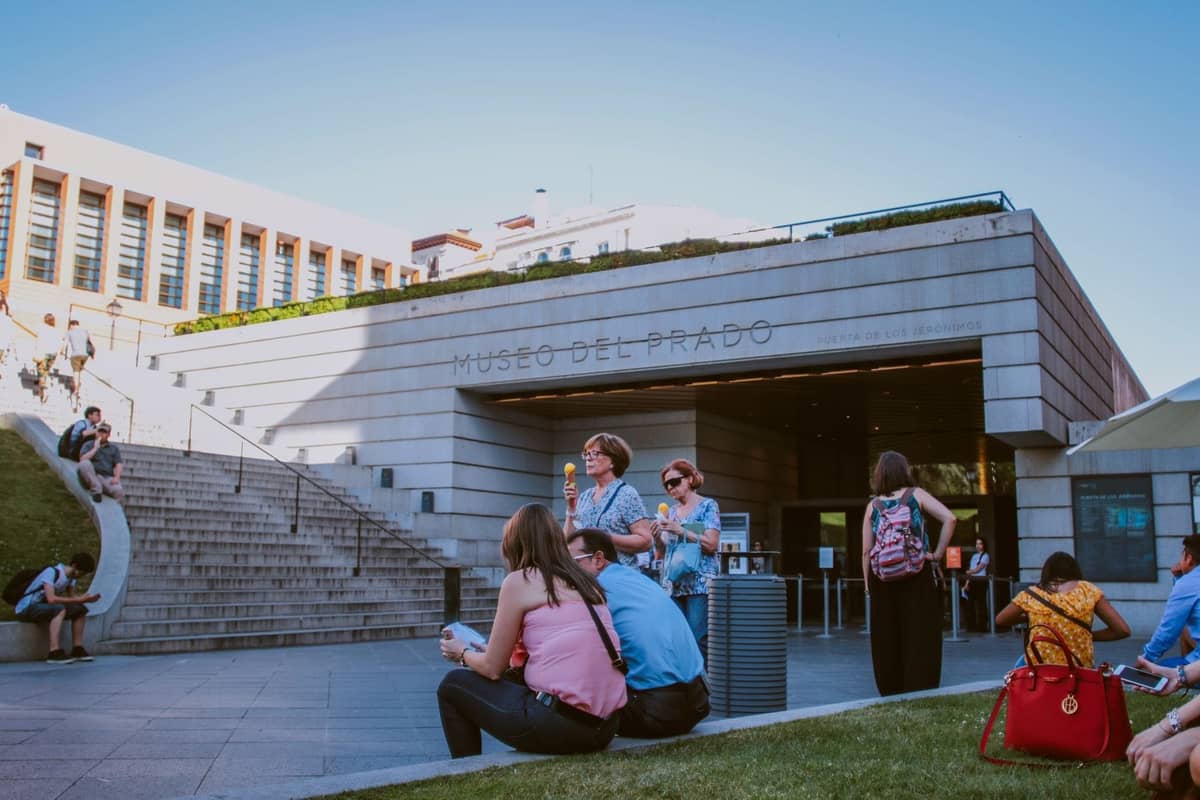 Persone, tra cui alcuni bambini, sedute e riunite all'esterno dell'ingresso moderno del Museo del Prado di Madrid in una giornata di sole.