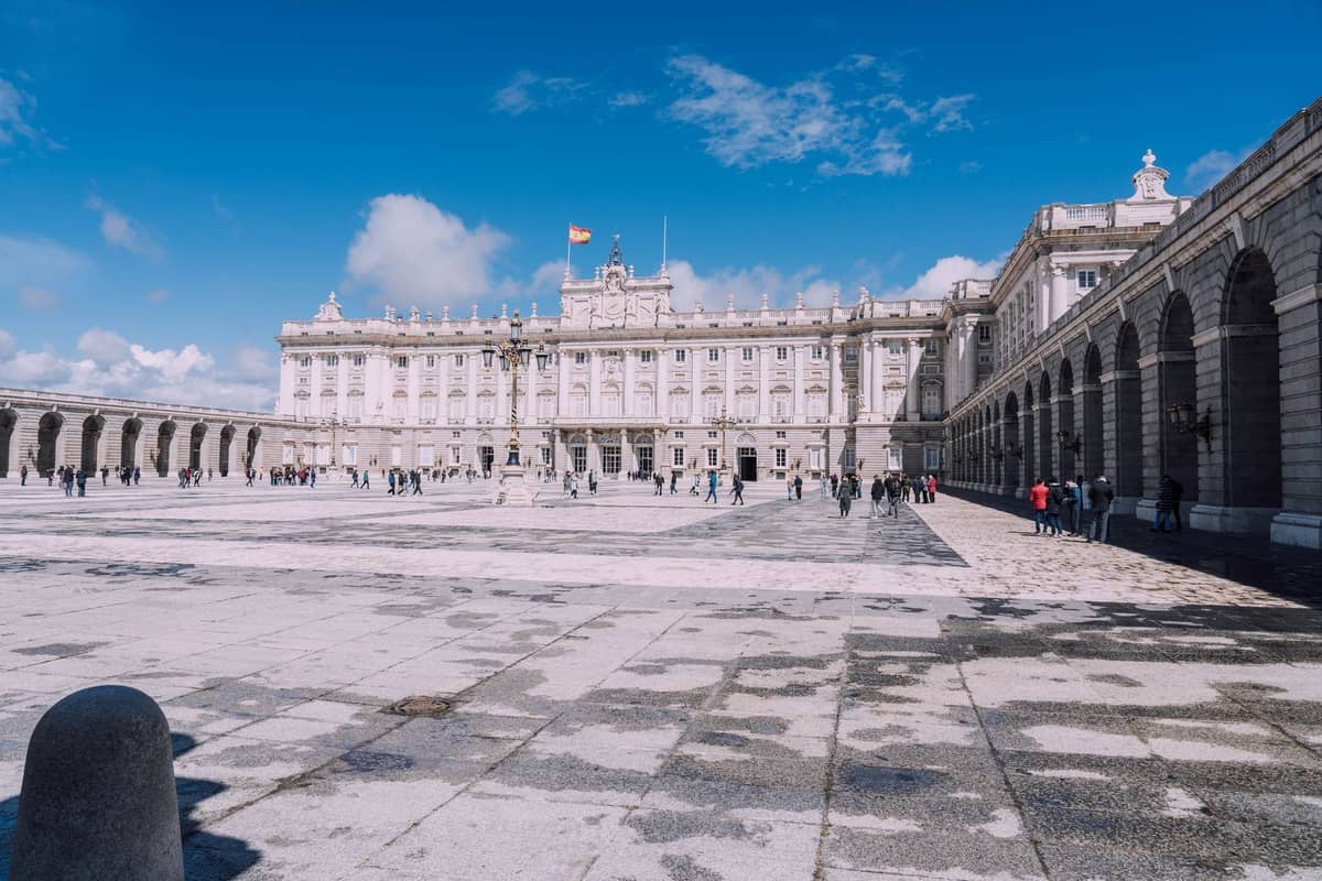 Vista ampia del Palazzo Reale di Madrid con persone che camminano nel grande cortile aperto sotto un cielo azzurro luminoso.