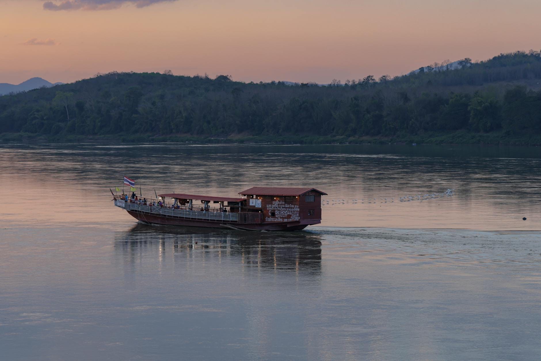 Una lancha tradicional con cubierta techada navega por un río tranquilo al atardecer, con colinas boscosas al fondo.