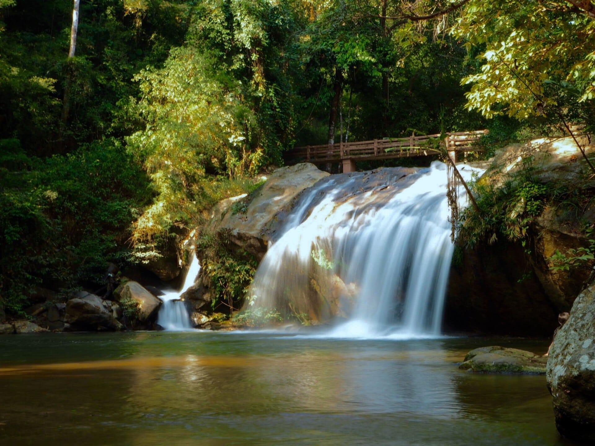 La cascada Mae Sa cae sobre grandes rocas bajo un puente de madera, rodeada de un frondoso bosque verde en el valle de Mae Rim.