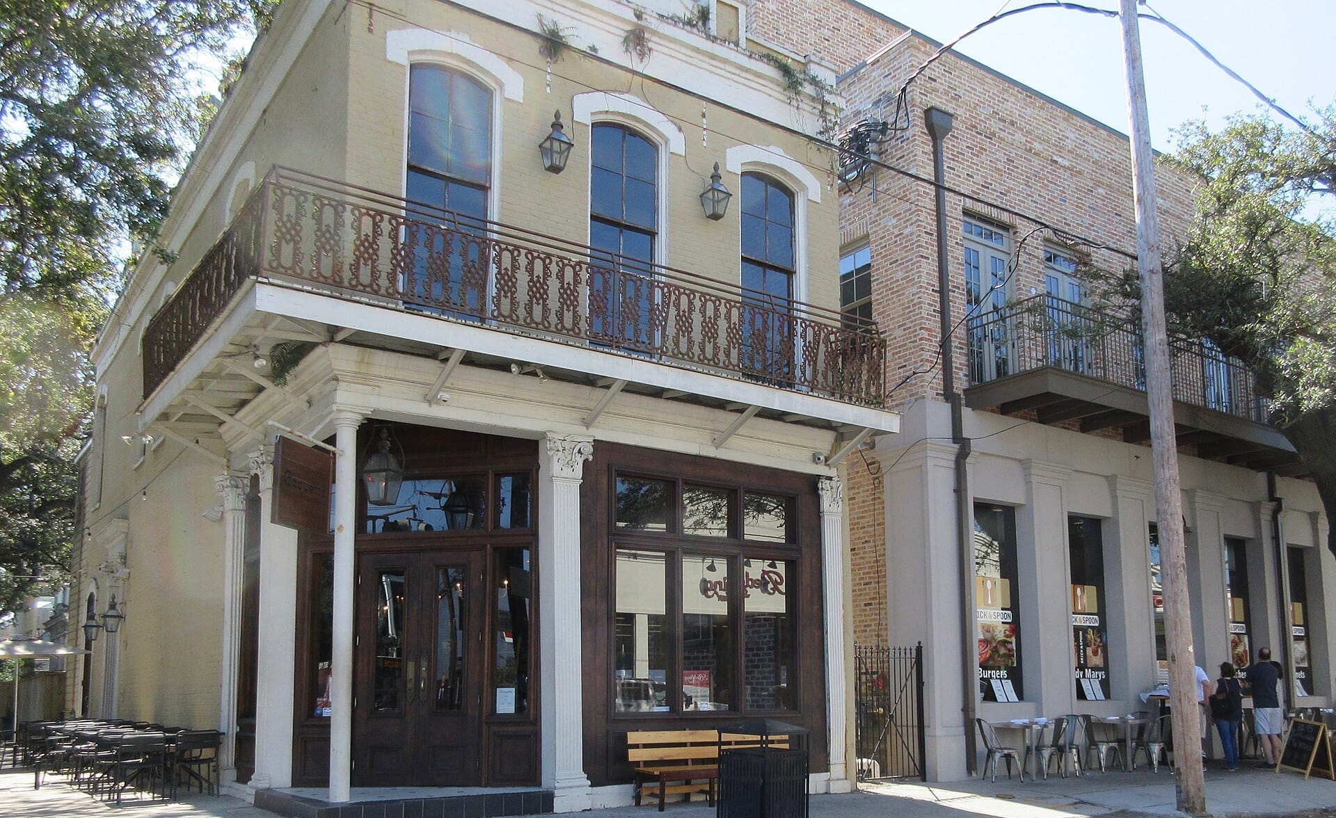 Storici edifici in mattoni con balconi e posti a sedere sul marciapiede su Magazine Street a New Orleans, ombreggiati da alberi frondosi in una giornata di sole.