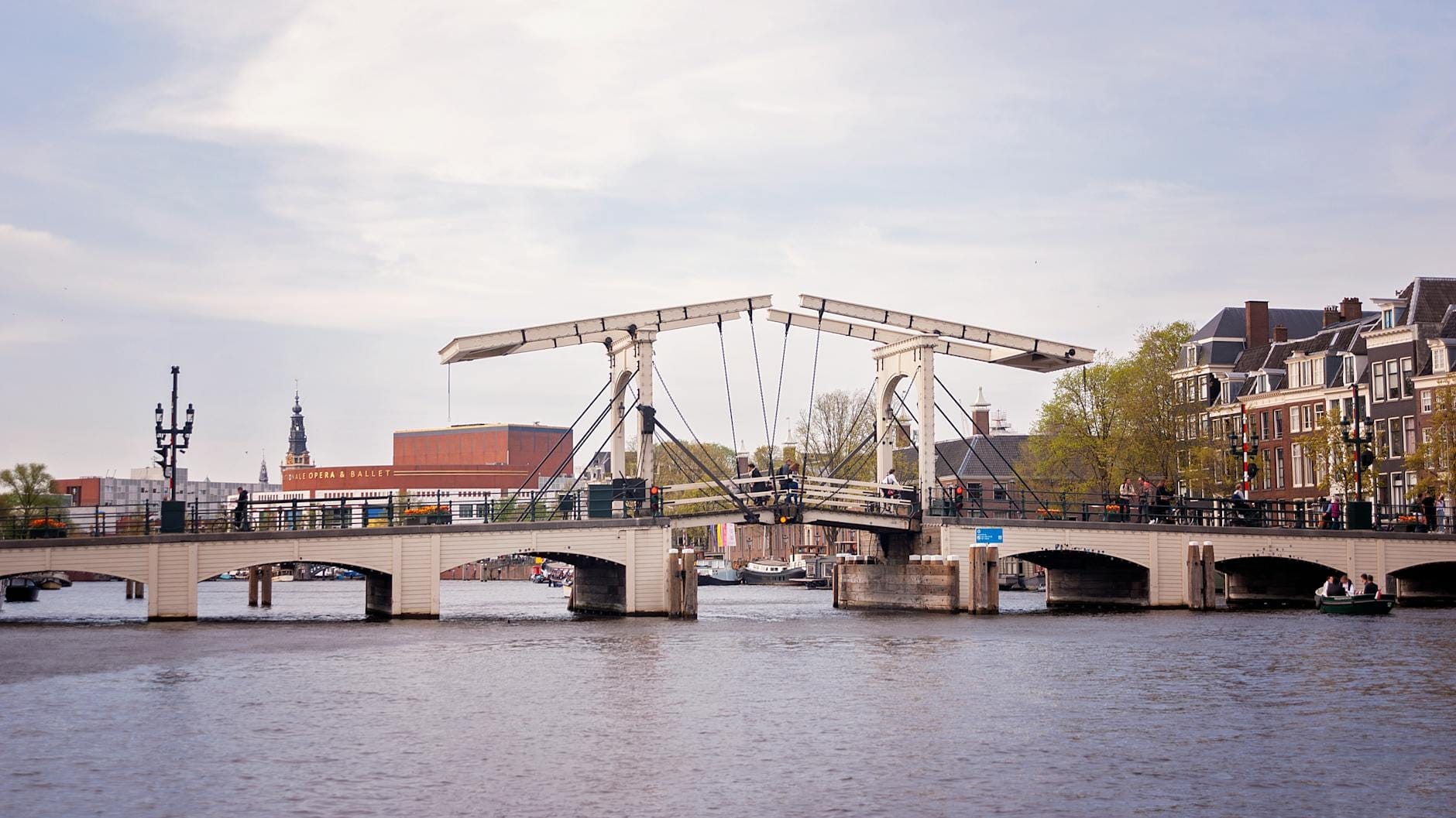 Vista panorámica del puente levadizo blanco Magere Brug sobre el río Amstel en Ámsterdam, rodeado de edificios y árboles bajo un cielo parcialmente nublado.