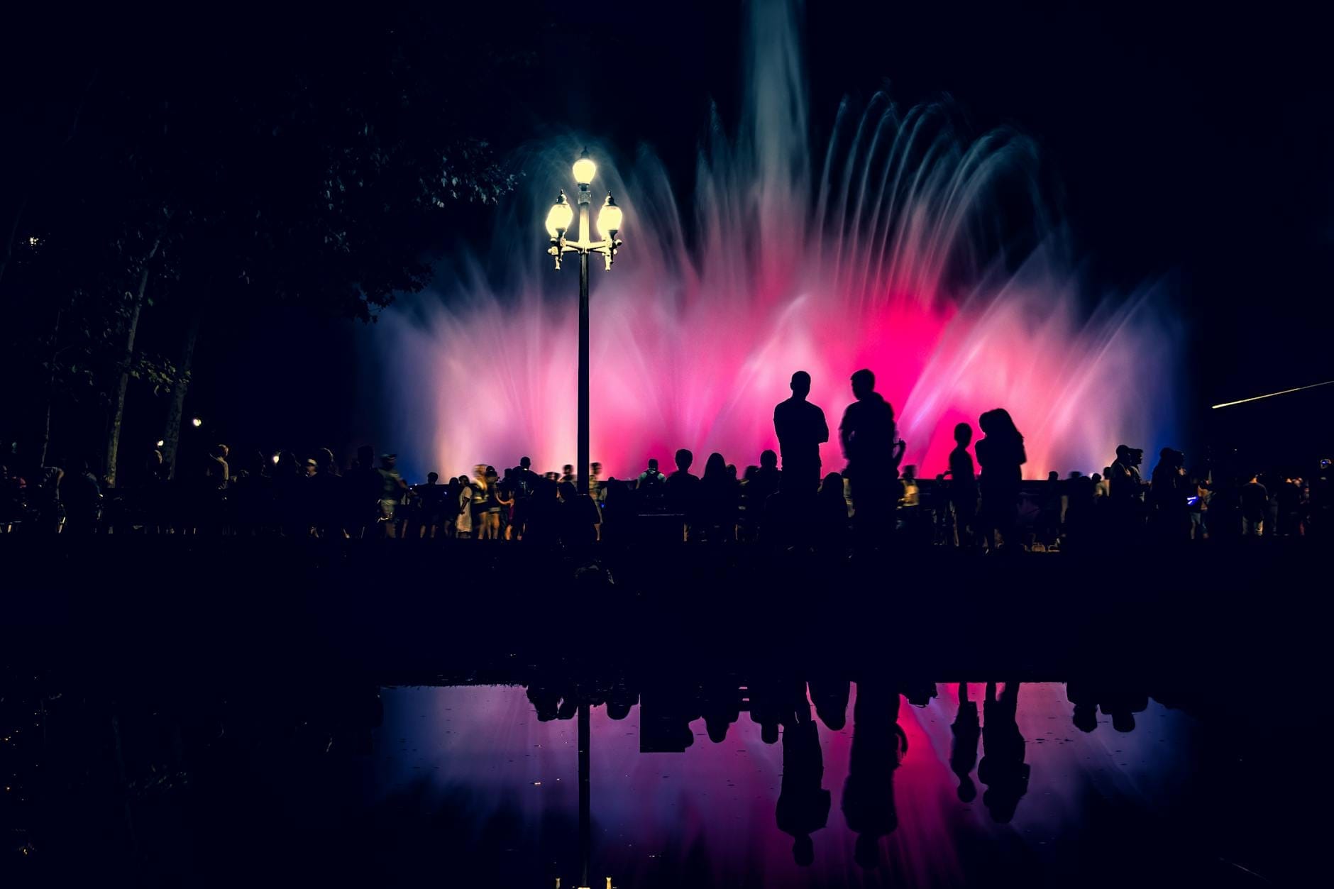 Large crowd silhouetted in front of the illuminated Font Màgica de Montjuïc at night with colorful water jets and reflections, creating a festive atmosphere.