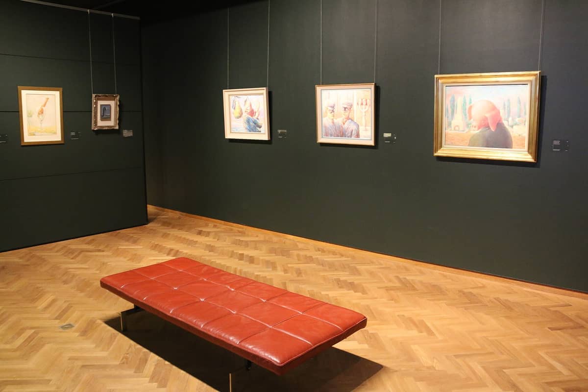 Galeria interior do Museu Magritte de Bruxelas com pinturas emolduradas em paredes verde-escuro e um banco de couro marrom sobre piso de parquê.