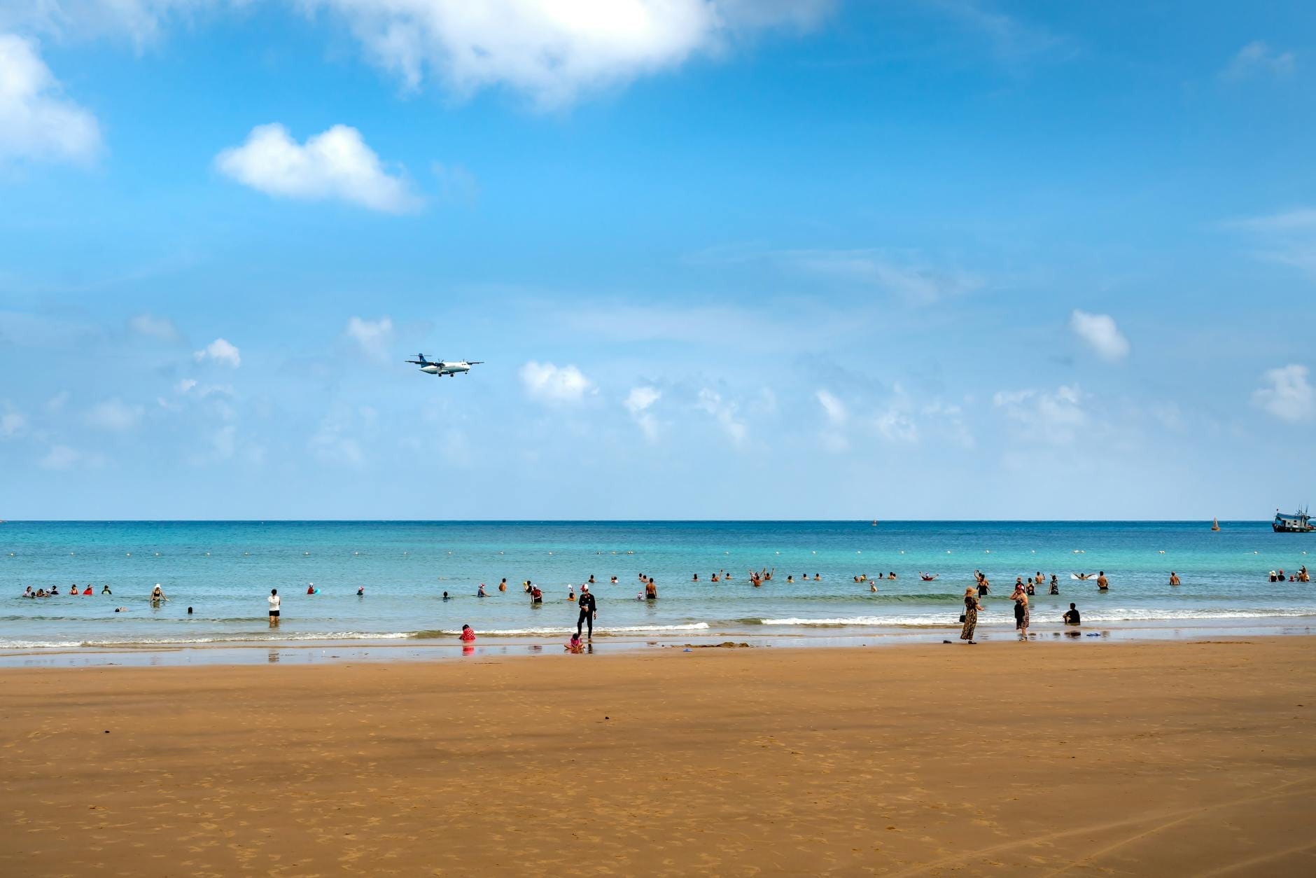 Wide sandy beach with clear blue water, people swimming and relaxing, and a plane flying low in the sky under bright clouds.