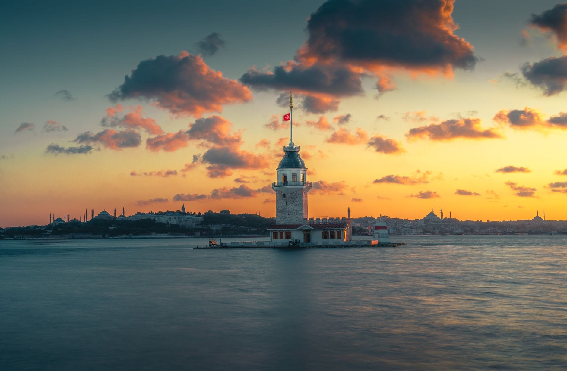 La Torre della Fanciulla si erge solitaria sul suo isolotto nel Bosforo al tramonto, con un cielo spettacolare e lo skyline di Istanbul sullo sfondo.