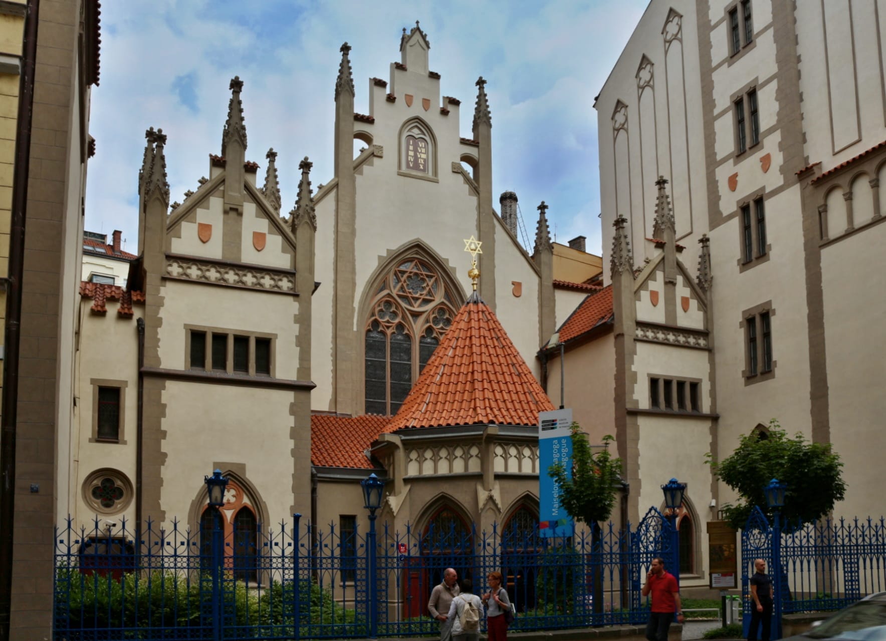 Synagoga Majzla w Pradze z ostrą neogotycką fasadą, łukowymi oknami, czerwonym dachówkowym dachem i zwiedzającymi przy wejściu.
