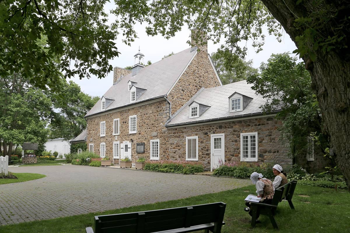 Blick auf die Maison Saint-Gabriel, ein historisches Steinbauernhaus in Montréal, mit Bäumen und kostümierten Führern, die im Garten auf Bänken sitzen.