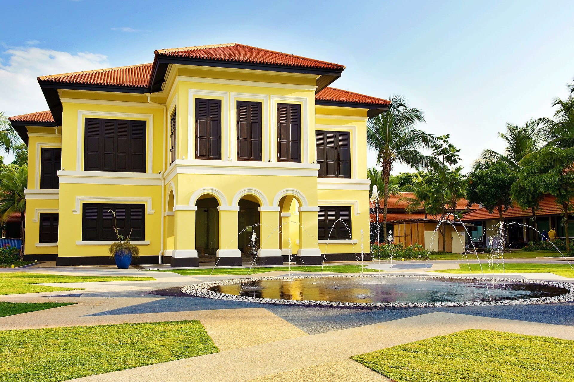 Malay Heritage Centre ในสิงคโปร์ อาคารอาณานิคมสีเหลืองสดใสพร้อมบานเกล็ดสีเข้ม ตั้งอยู่เบื้องหลังน้ำพุทรงกลมและสวนที่ตัดแต่งอย่างเรียบร้อยภายใต้ท้องฟ้าแจ่มใส