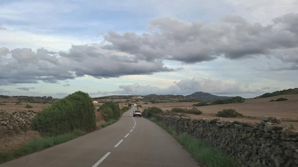 Con đường nông thôn hẹp ở Mallorca được flanked by những bức tường đá và cánh đồng, với một chiếc ô tô đang lái về phía những ngọn đồi xa dưới bầu trời nhiều mây.