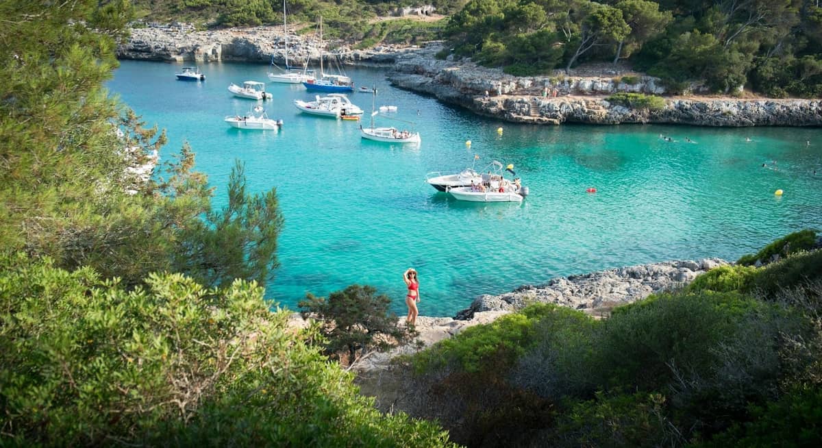 Cala turquesa apartada en Mallorca con barcas ancladas y árboles enmarcando acantilados rocosos, destacando un destino accesible solo por mar.