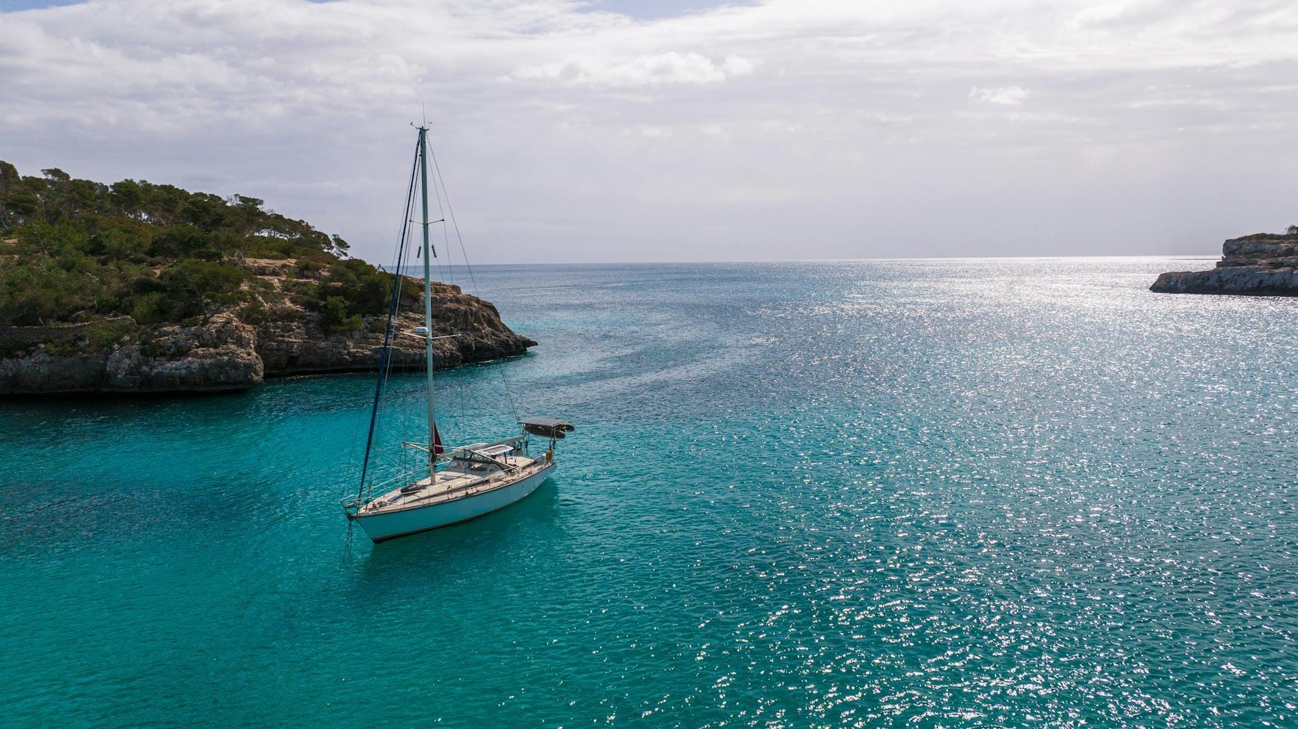 Um veleiro ancorado em uma deslumbrante enseada de águas turquesa, emoldurado por rochas e pinheiros sob um céu azul intenso, evocando a serenidade da navegação em Maiorca.
