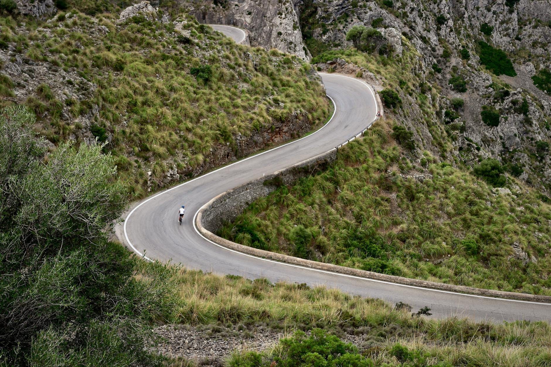 Un ciclista solitario affronta un tornante spettacolare su una strada di montagna sinuosa, circondata da pareti rocciose e prati verdi nella Serra de Tramuntana.