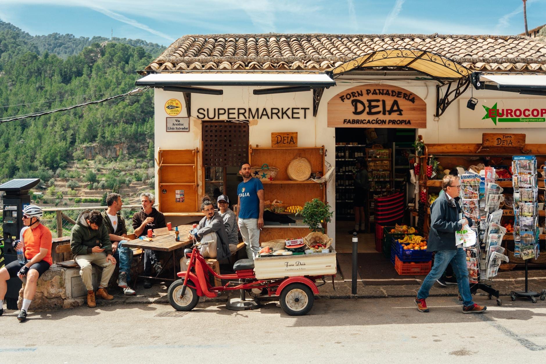 Uma animada padaria e feira de aldeia mallorquina com pessoas sentadas do lado de fora, rodeadas de produtos locais, montanhas ao fundo, luz do sol e uma atmosfera mediterrânea descontraída.