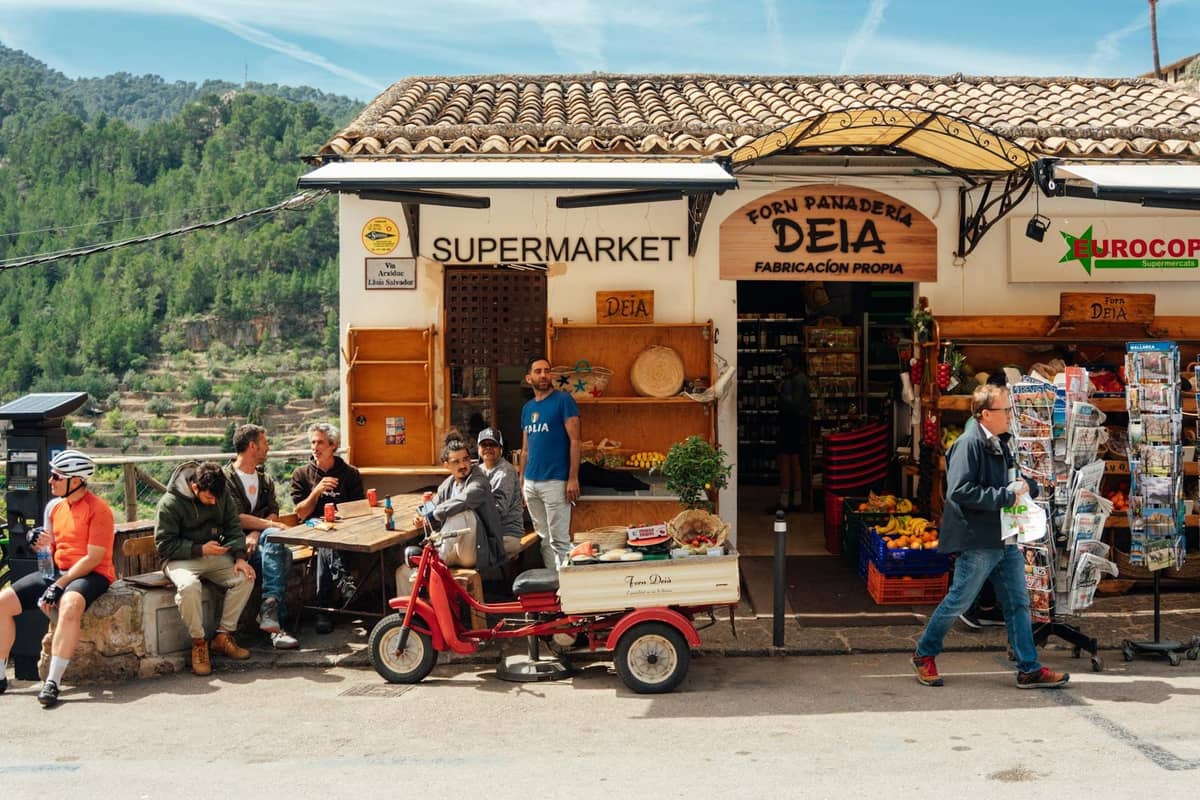 Straßenszene vor einer Bäckerei und einem Supermarkt in Deià, Mallorca, mit Menschen an Tischen im Freien und sichtbaren lokalen Produkten.